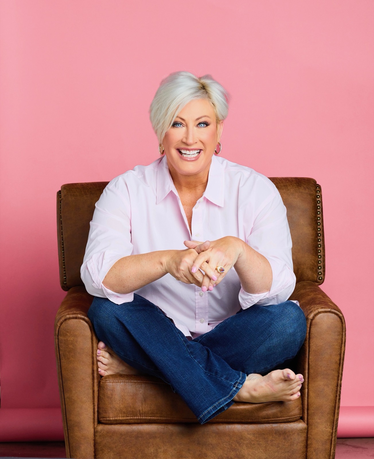 Kim Gravel sitting cross-legged in a leather armchair, laughing, against a pink backdrop